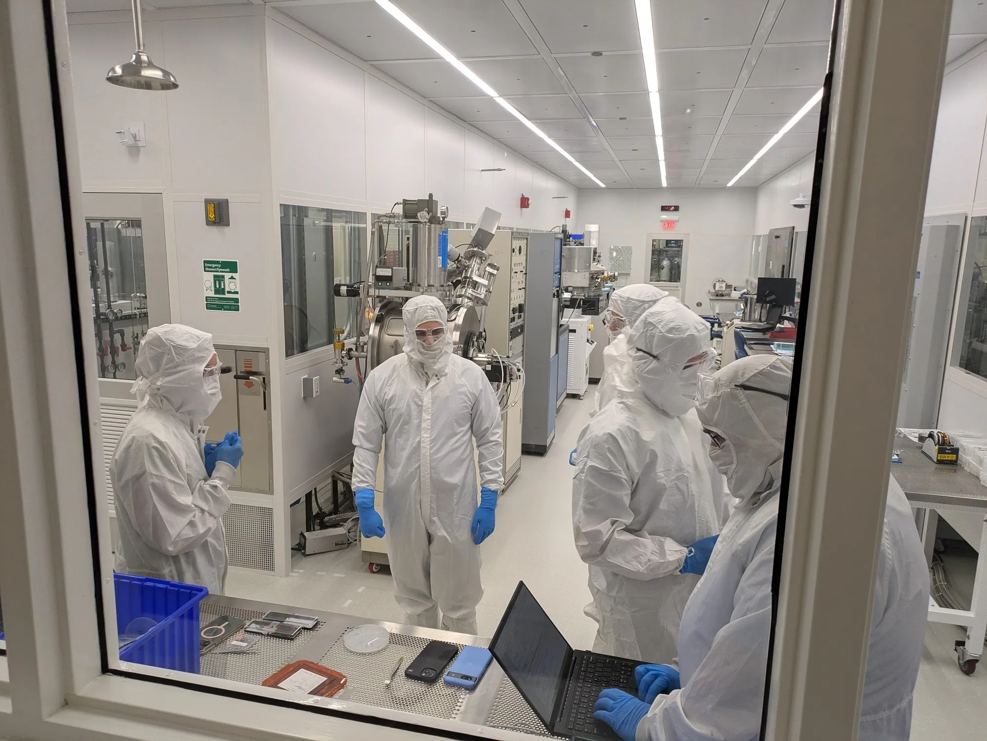Five gowned Advanced Optronics engineers at work in the nanofabrication cleanroom, seen through the bay window — one at a deposition tool, three in conversation, one at a laptop in the foreground.