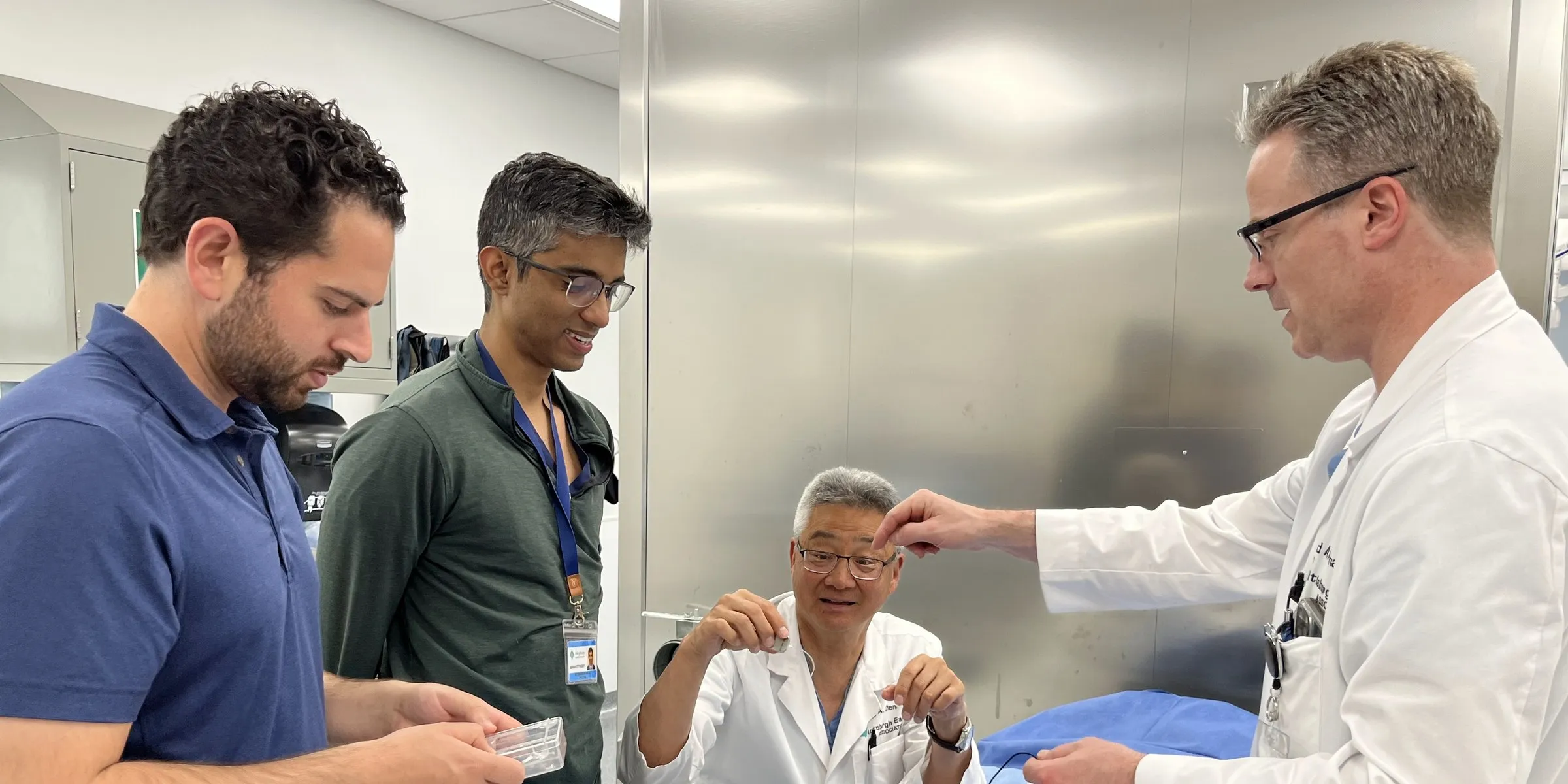 Two Advanced Optronics engineers and two cochlear implant surgeons in conversation around a work table in a hospital procedure room, one surgeon gesturing behind his ear.