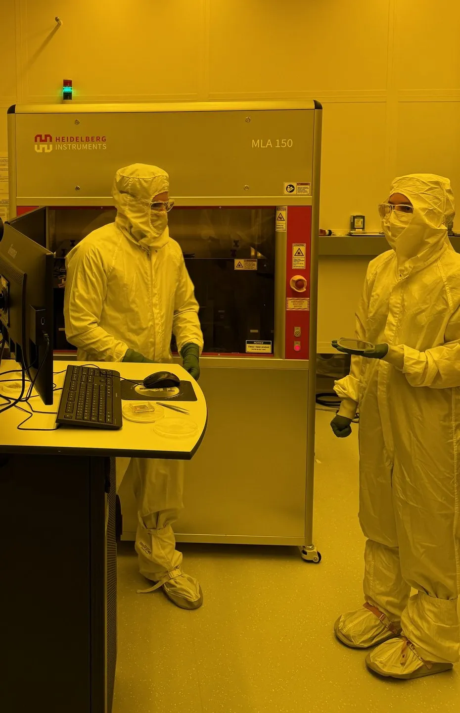 Two gowned engineers at a maskless lithography aligner in the cleanroom, one holding a wafer, under yellow light.