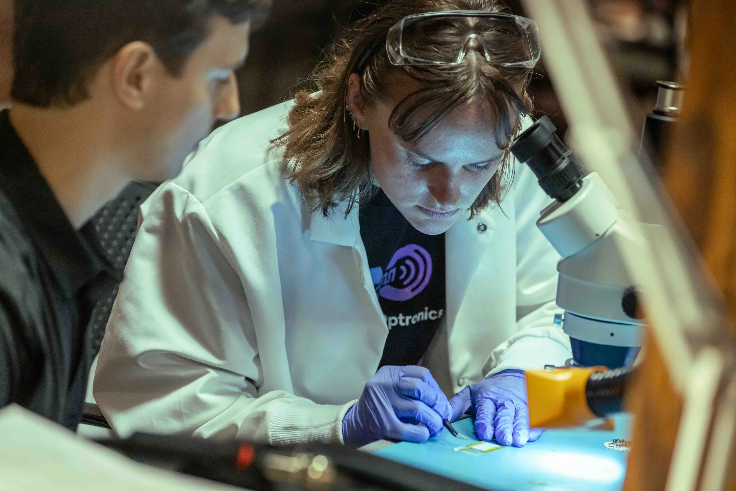 An engineer in a lab coat and safety goggles works on a device beneath a stereo microscope while a colleague looks on.