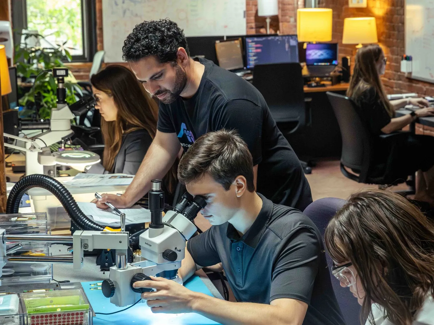 The Advanced Optronics bench in the brick-walled office: Jay Reddy standing in a black AO tee looking over a colleague working at a stereo microscope, a third engineer at a scope to the left and a fourth in a white coat and safety glasses handling a device at the right.